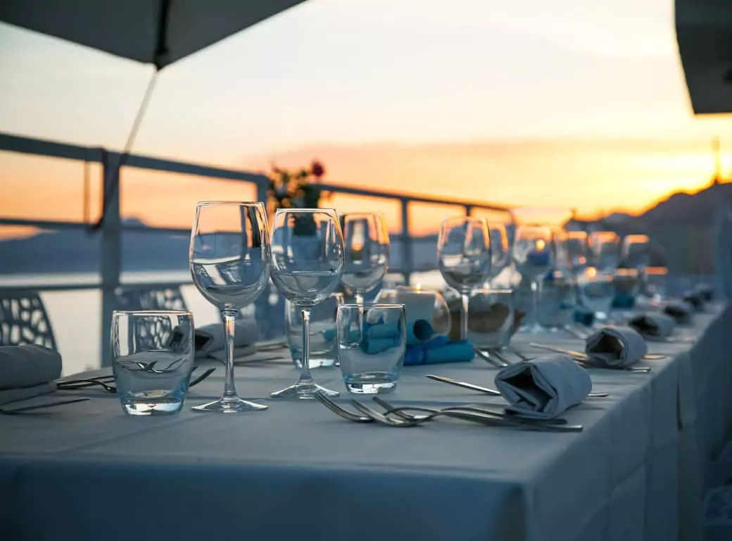 A beautifully set outdoor dining table with elegant glassware and neatly folded napkins, overlooking a stunning sunset by the water. Discover must-try dining experiences with Ambl.