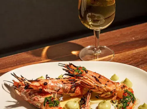 A plate of grilled prawns garnished with fresh herbs and chili, served alongside dollops of a green sauce, paired with a chilled glass of white wine on a wooden table. Discover vibrant dining in Shoreditch with Ambl.