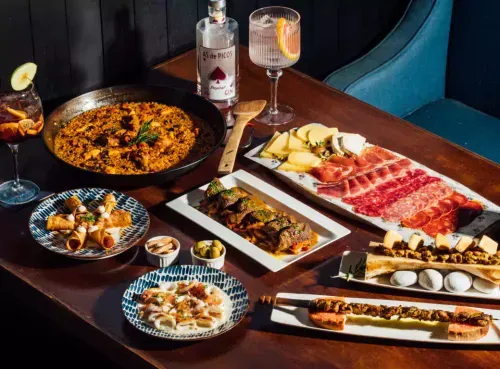 A beautifully arranged spread of Spanish cuisine, featuring a rich paella, an assortment of cured meats and cheeses, crispy tapas, and flavorful grilled skewers. Two elegant gin cocktails and small plates complete the inviting setup. Discover authentic Spanish dining with Ambl.