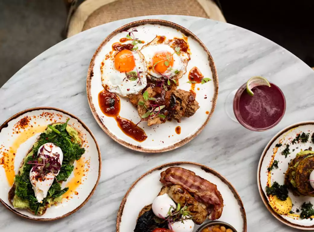 A beautifully plated brunch spread, featuring dishes like fried eggs with crispy fried chicken, avocado toast with poached eggs, a full English breakfast, and a savory stack with hollandaise. Accompanied by a vibrant berry cocktail, this setup is perfect for a business breakfast or weekend brunch. Discover top breakfast spots with Ambl.