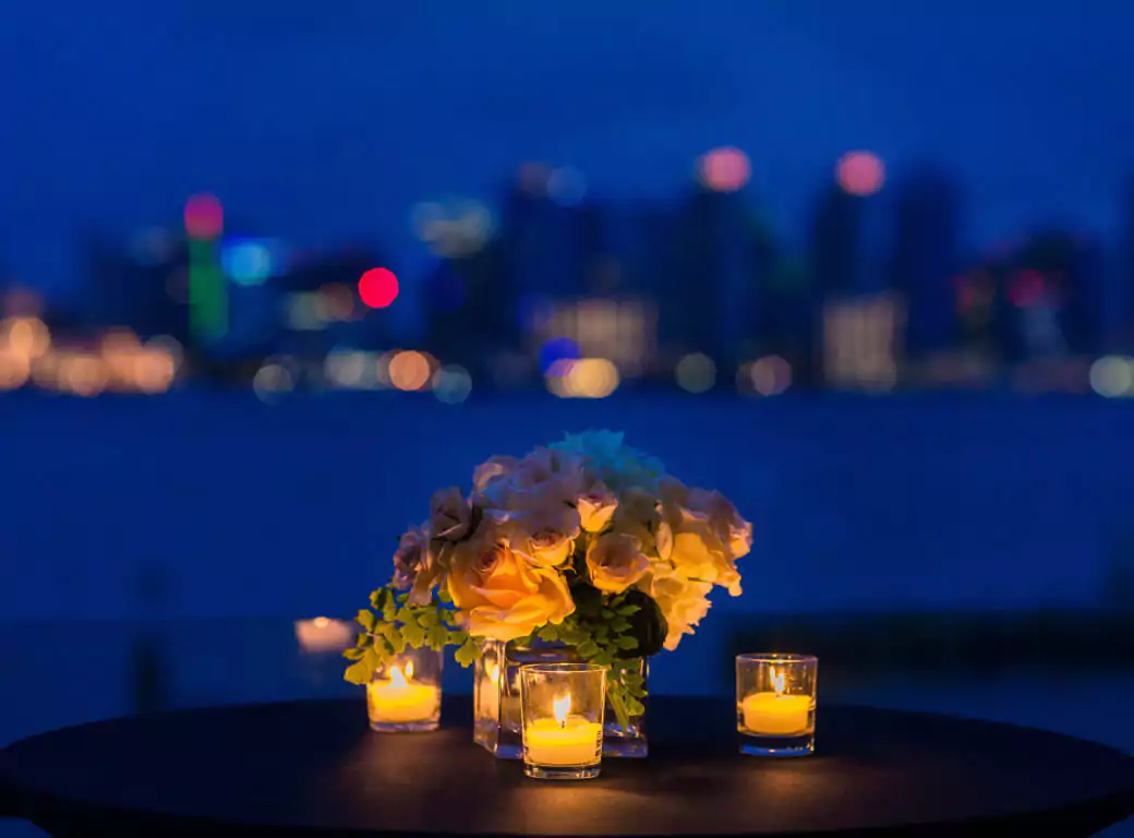 A romantic candlelit table set against the shimmering city skyline at night. Elegant yellow roses and glowing candles create an intimate atmosphere, perfect for a waterfront dinner or special occasion. Discover the most picturesque dining spots with Ambl.