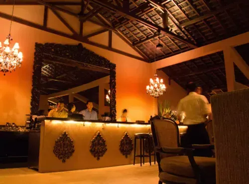 A luxurious Indian restaurant with warm amber lighting, intricate carved décor, and an ornate mirror reflecting the stylish bar. Chandeliers cast a soft glow over the elegant seating and attentive staff. Experience the rich flavors and opulence of Indian cuisine with Ambl.