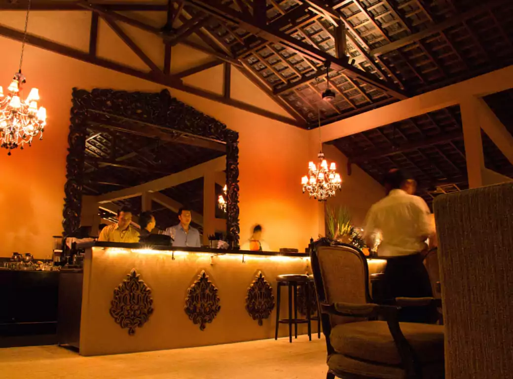 A luxurious Indian restaurant with warm amber lighting, intricate carved décor, and an ornate mirror reflecting the stylish bar. Chandeliers cast a soft glow over the elegant seating and attentive staff. Experience the rich flavors and opulence of Indian cuisine with Ambl.