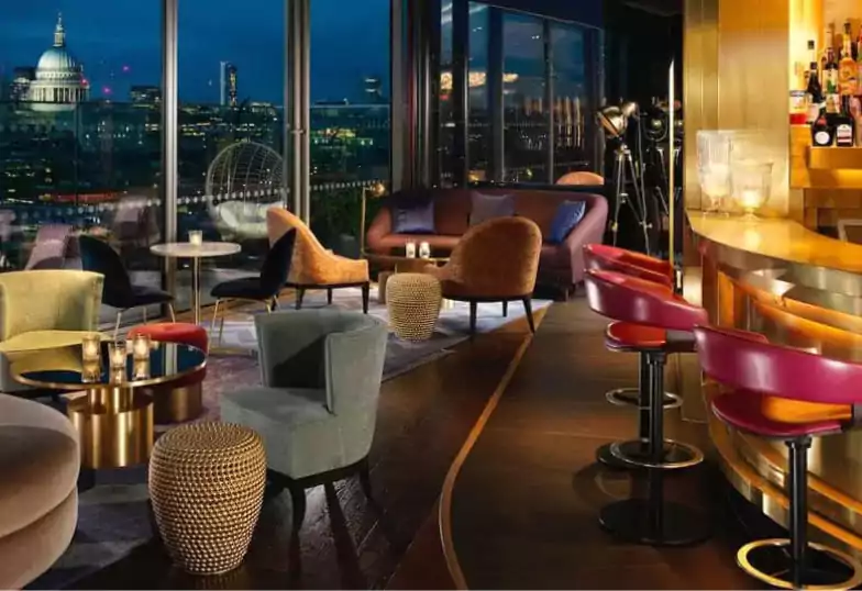 A luxurious rooftop bar with plush seating, golden accents, and panoramic views of London’s skyline at night. Candles flicker on tables, creating an inviting atmosphere for a stylish evening out. Find more bars like this on Ambl.