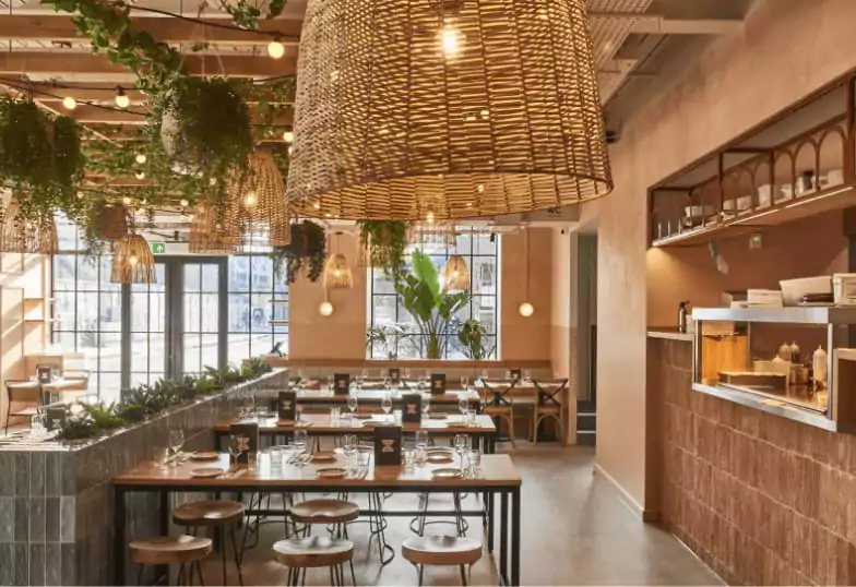 A warm and rustic Mediterranean-inspired eatery at Brother Marcus, adorned with hanging plants, wicker light fixtures, and a welcoming ambiance. Find stylish spots like this with Ambl.