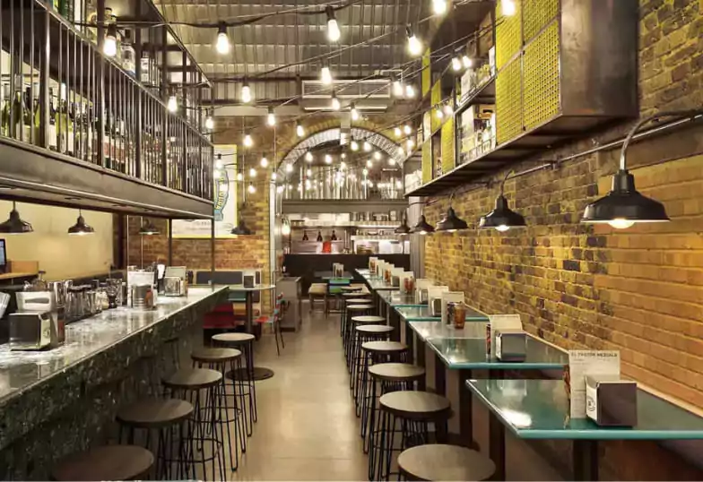 A rustic and industrial-chic taqueria set under brick archways with string lights, long communal tables, and a welcoming ambiance with Ambl.