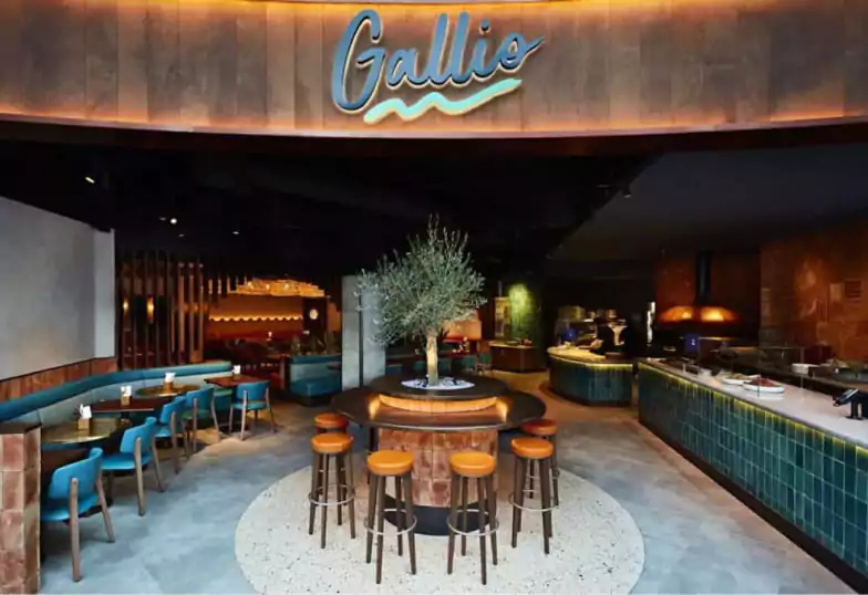 A stylish Mediterranean eatery with teal booths, wooden bar seating, and a central olive tree, blending contemporary design with rustic charm. A perfect spot for sharing plates and cocktails. Discover more Mediterranean-inspired spaces on Ambl.