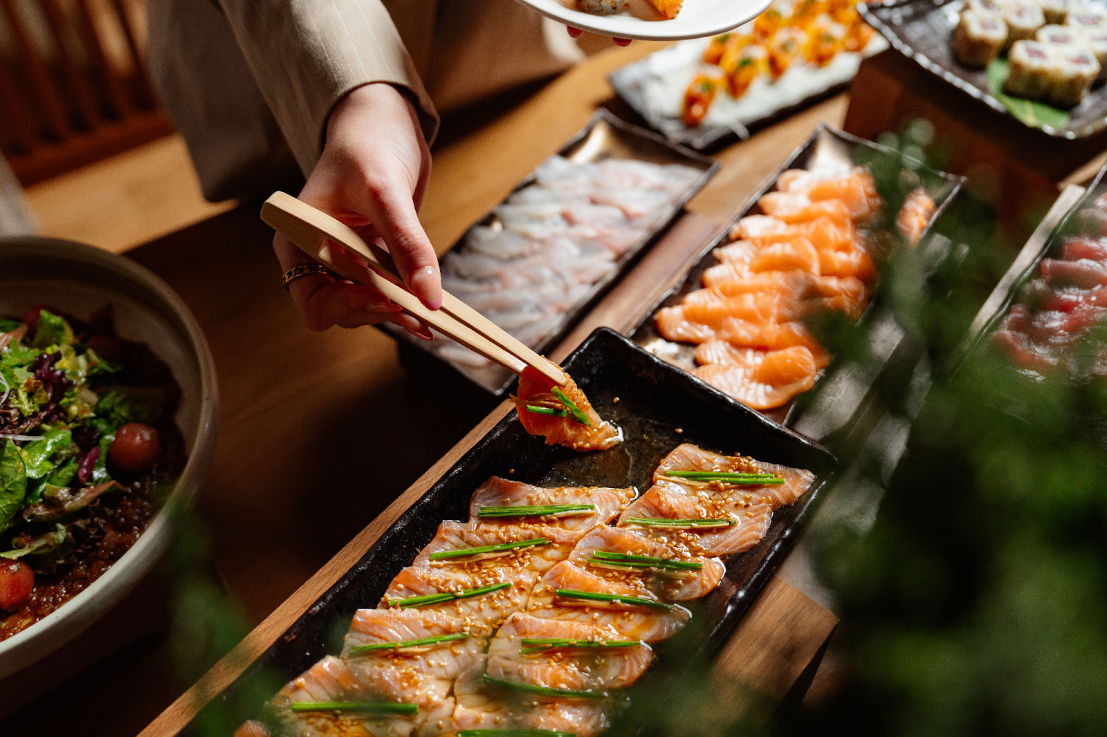 A person holding chopsticks above a tray filled with various sushi pieces, ready to pick one up.