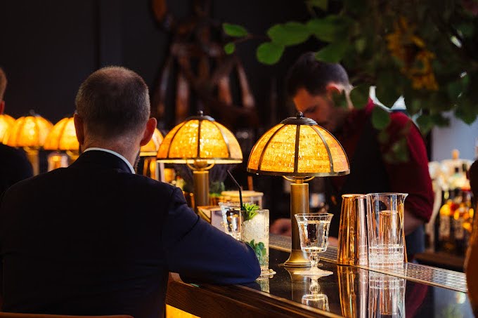 A man seated at a bar, holding a glass of red wine, with a relaxed expression on his face.