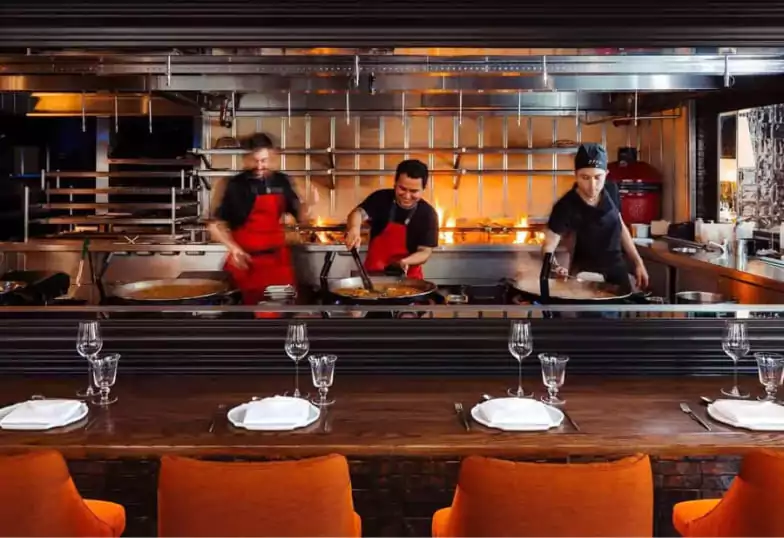 A vibrant open kitchen where chefs prepare authentic paella over a fire, set in a stylish, modern space. Find the best Spanish cuisine with Ambl.