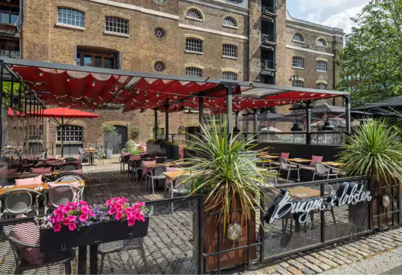 A spacious outdoor dining area at Burger & Lobster, featuring red canopy shades, lush greenery, and cozy seating. Discover al fresco dining with Ambl.