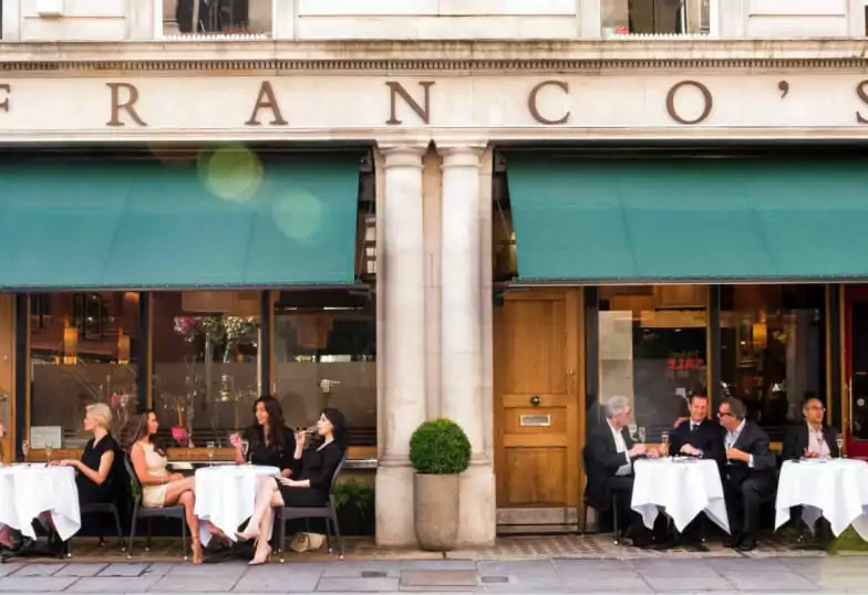 A refined European café with green awnings, white-clothed tables, and a classic storefront, offering an elegant outdoor dining experience. A timeless choice for fine dining. Find more classic venues on Ambl.