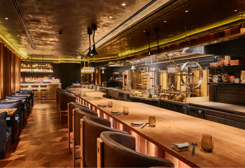 An immersive dining space with a dramatic open kitchen, a sleek bar, and an elegant wood-accented interior, showcasing culinary artistry. Explore more unique dining concepts on Ambl.