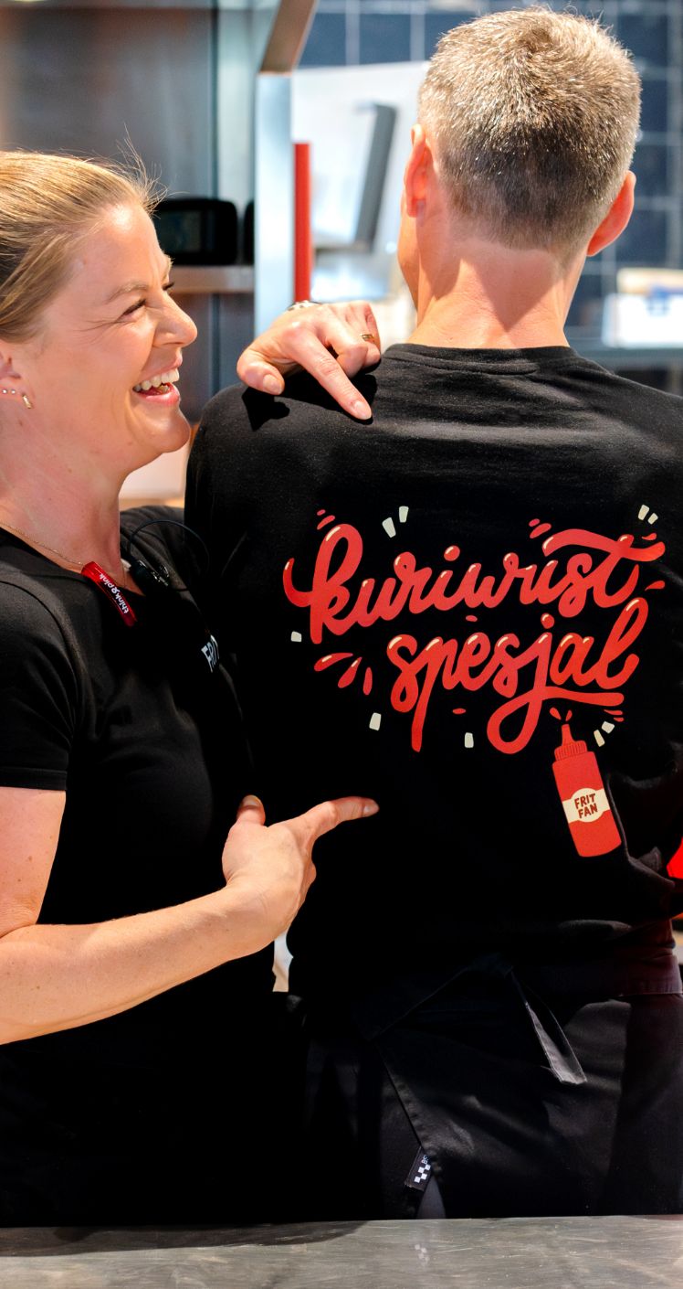 Vrouw lacht en wijst naar T-shirt met tekst "Currywurst Speciaal" in restaurant.