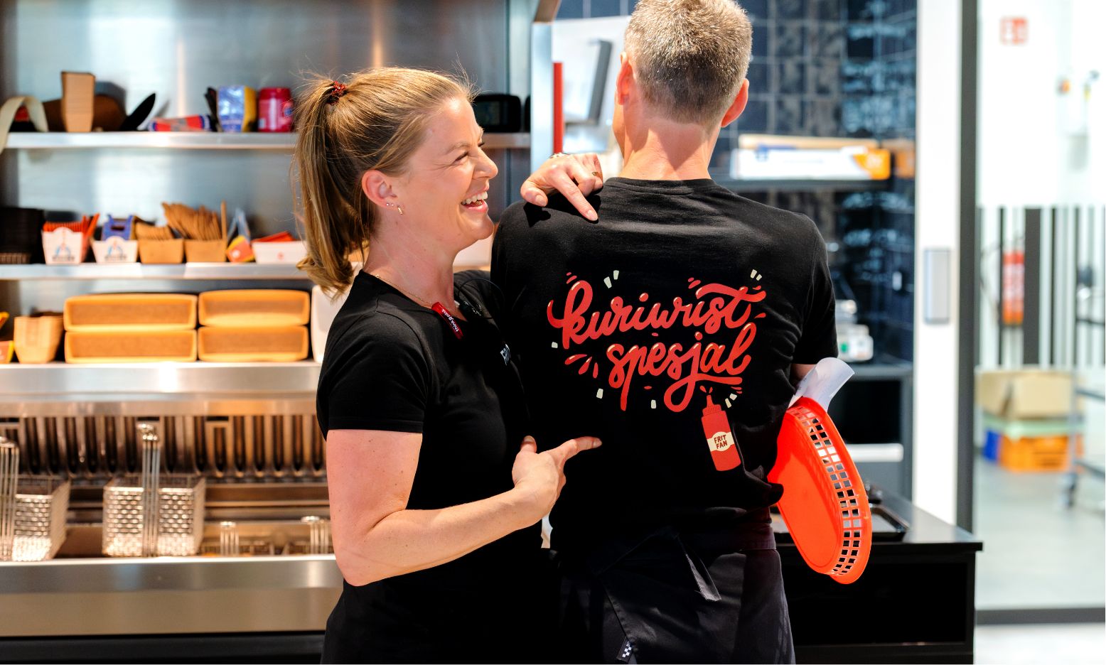 Vrolijke vrouw in snackbar wijst naar man met "Kiriwurst Speciaal" T-shirt.