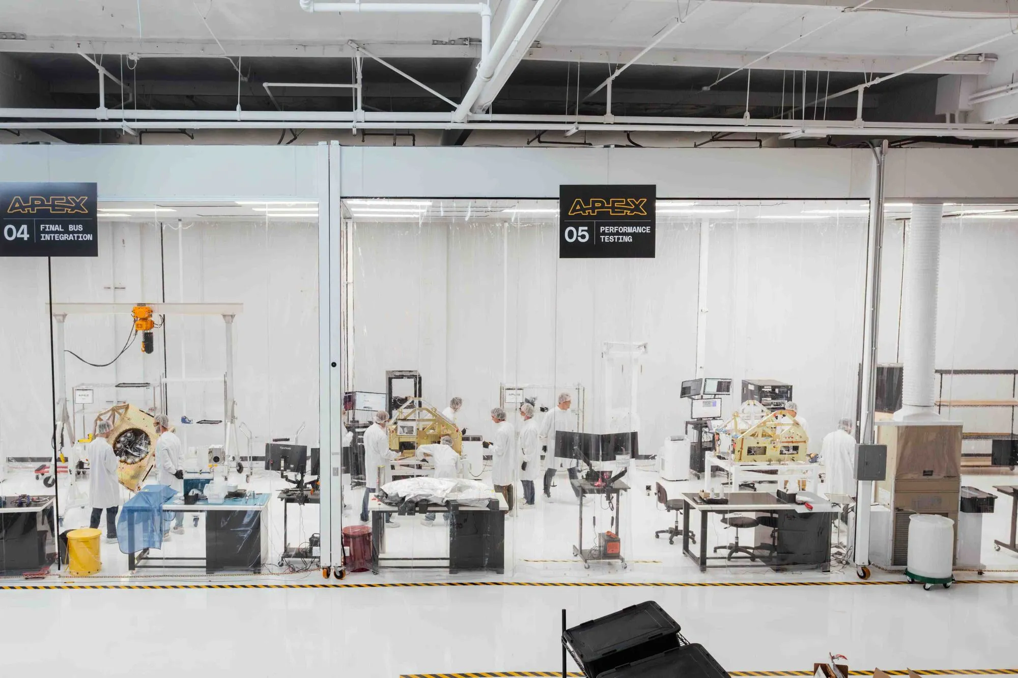 Laboratory workspace with multiple technicians in white coats and hairnets working on aerospace components behind glass partitions labeled 'Final Bus Integration' and 'Performance Testing'.