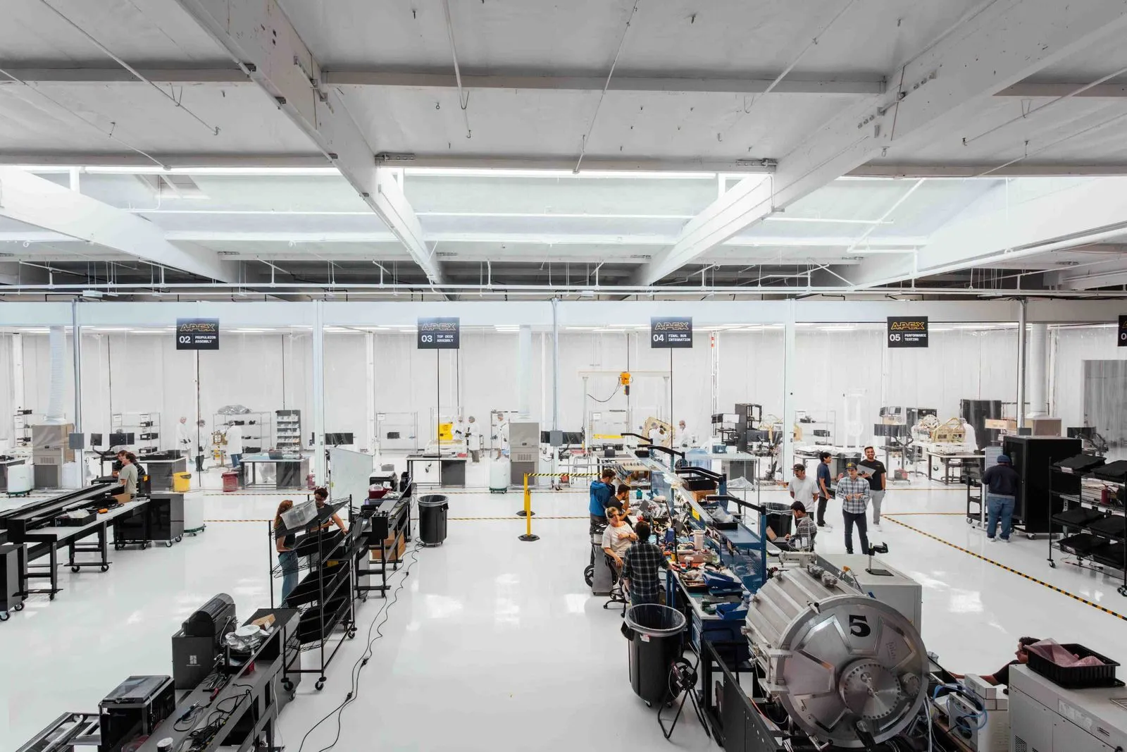Bright, spacious factory floor with workers assembling and inspecting equipment at organized stations under skylights.