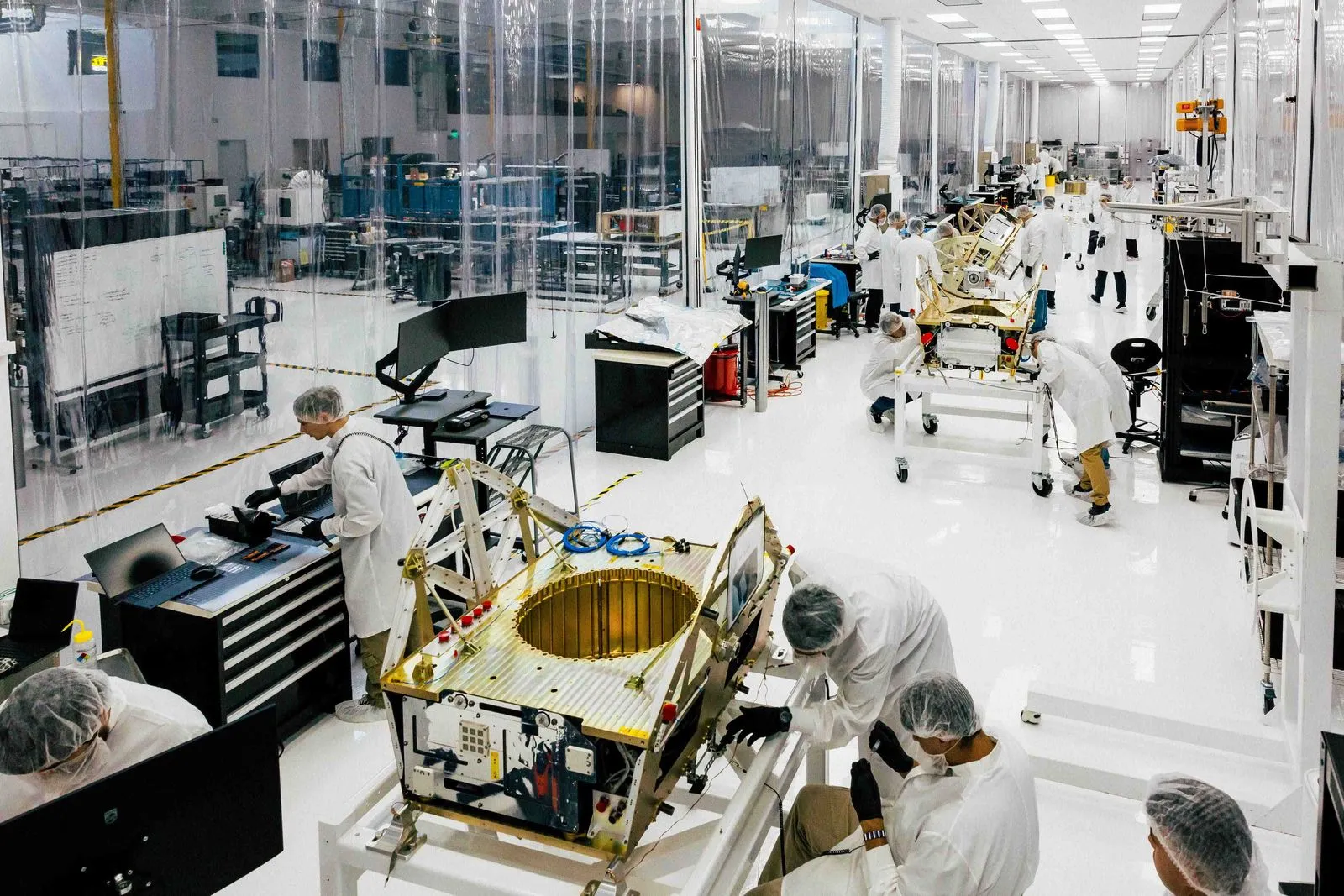 Technicians in white lab coats and hairnets assembling spacecraft components in a clean, white manufacturing facility.
