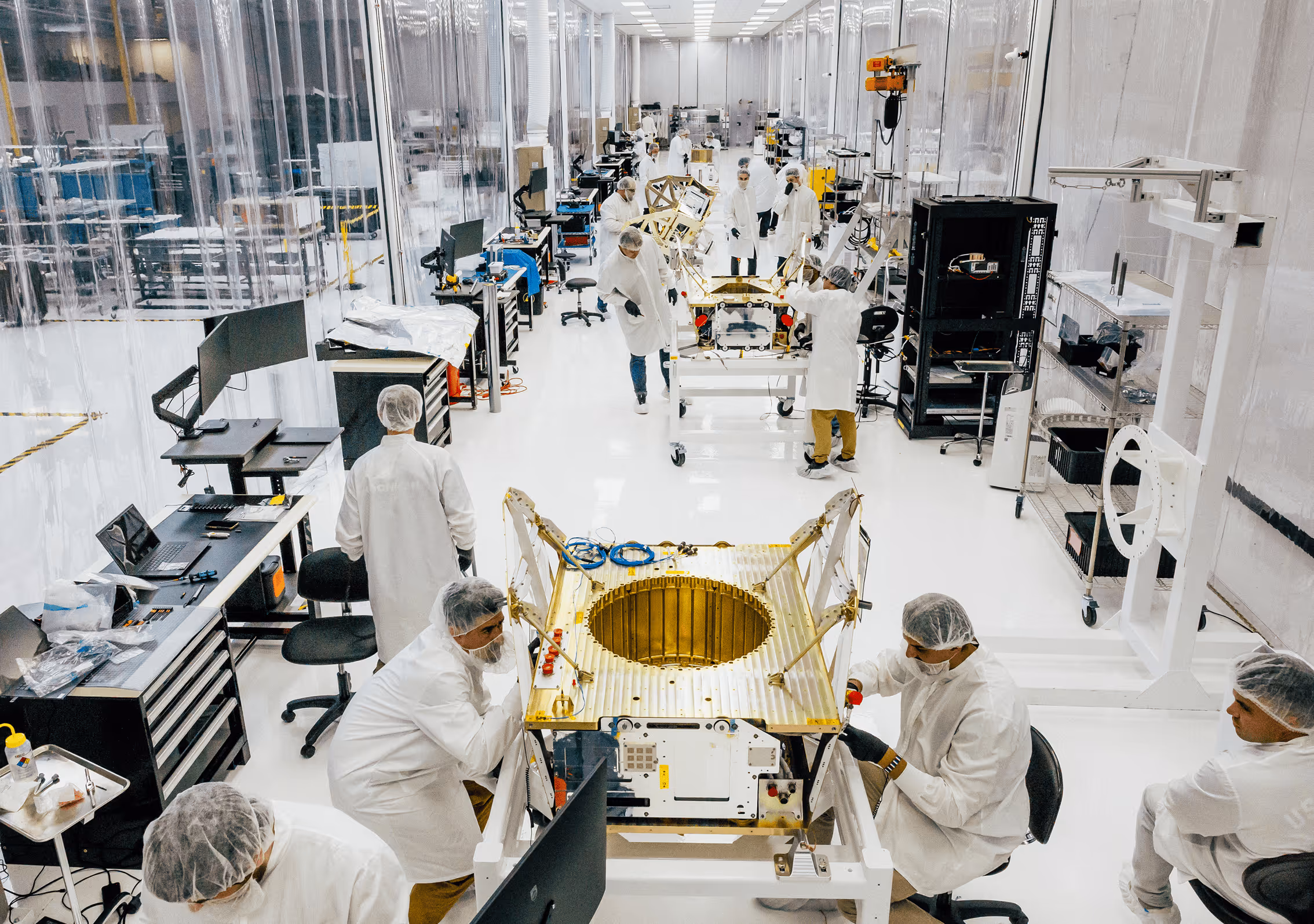Engineers in white lab coats and hairnets assemble and inspect gold-colored spacecraft components in a clean, high-tech factory environment.