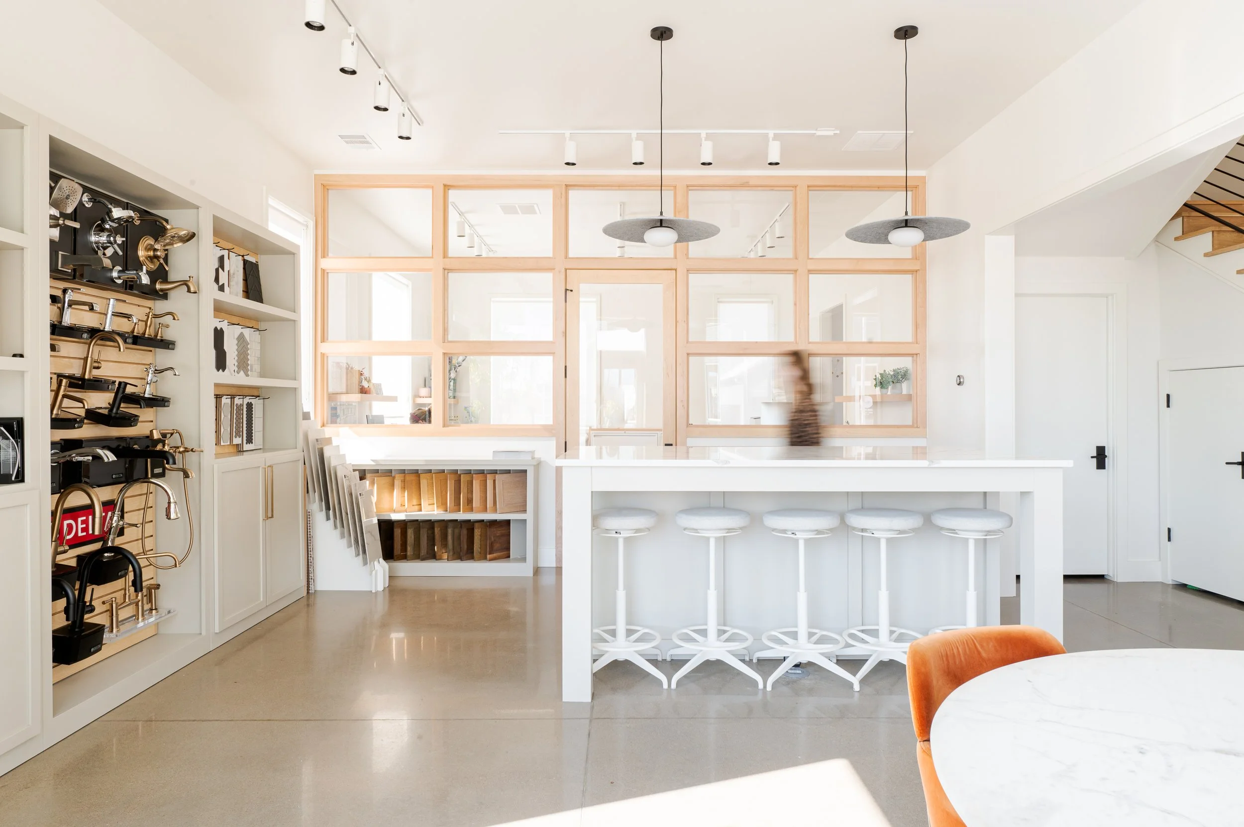 Modern white kitchen showroom with a large island, five white bar stools, pendant lights, and wall displays of faucets and wood samples.