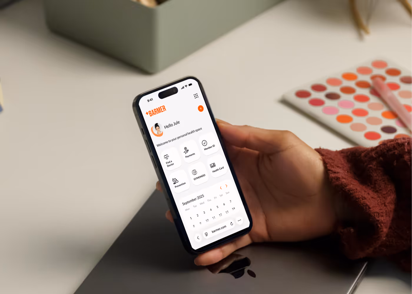 Hand holding smartphone displaying calendar and profile view of an app, laptop and dotted notebook in background.