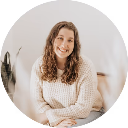 Woman with brown curly hair wearing a textured knit sweater sitting with hands resting on their lap in a softly lit room with a plant in the background.