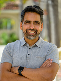 Smiling man with dark hair and beard wearing gray polo and smartwatch, arms crossed outdoors.