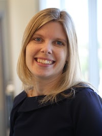 Smiling woman with straight blonde hair wearing a dark top, standing indoors near a window.