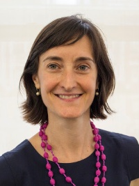 Smiling woman with short brown hair wearing a dark top and a long pink beaded necklace.