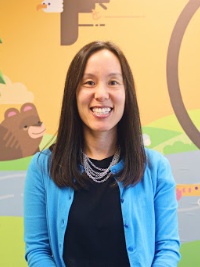 Smiling woman with long dark hair wearing a blue cardigan and black top, standing in front of a colorful mural.