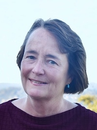 Smiling woman with short brown hair wearing a burgundy top and blue earrings outdoors.