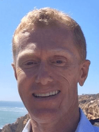 Smiling man with short light hair outdoors near water and rocky shore under a clear blue sky.