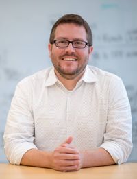 Smiling man with glasses wearing a white shirt sitting at a table with blurred whiteboard behind him.