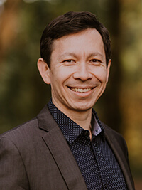 Smiling man with dark hair wearing a dark blazer and patterned shirt outdoors.