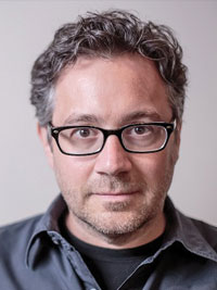 Man with curly gray hair and glasses wearing a dark collared shirt against a plain background.