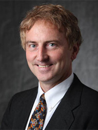 Smiling man with curly light brown hair wearing a suit, white shirt, and patterned tie against gray background.