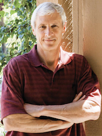 Man with gray hair wearing a maroon polo shirt stands with arms crossed near a beige wall and greenery.