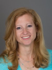 Smiling woman with blonde hair wearing a blue top and layered necklaces against a gray background.