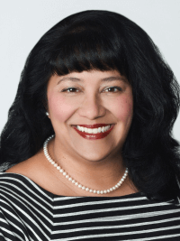 Smiling woman with black hair wearing a striped top and pearl necklace on a light background.