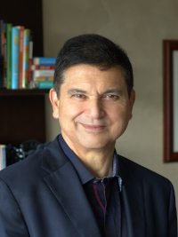 Man with dark hair wearing a navy blazer and striped shirt smiling indoors with bookshelves in background.