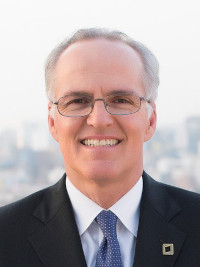 Smiling mature man with glasses wearing a suit and tie against a cityscape background.