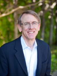Smiling man with glasses wearing a dark blazer and white shirt in an outdoor setting with green trees.