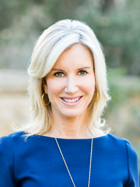 Smiling woman with blonde hair in a blue top standing outdoors with a blurred natural background.