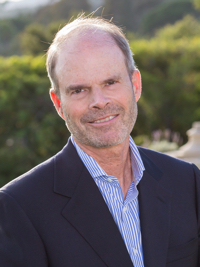 Smiling middle-aged man in a dark blazer and striped shirt outdoors with green foliage background.
