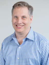 Smiling man wearing a blue checkered shirt against a plain light gray background.