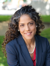 Smiling woman with curly hair wearing a navy blazer, red top, and pearl necklace outdoors.