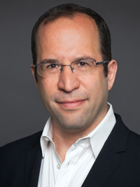 Man with glasses and short dark hair wearing a white shirt and black blazer on gray background.
