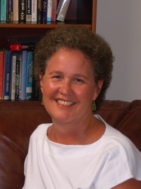 Smiling woman with short curly hair wearing a white shirt sitting in front of a bookshelf.