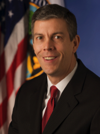 Man in suit with red tie smiling in front of U.S. flag and another flag on blue background.
