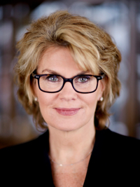 Portrait of a woman with blonde hair, glasses, and a black top smiling softly in a blurred background.